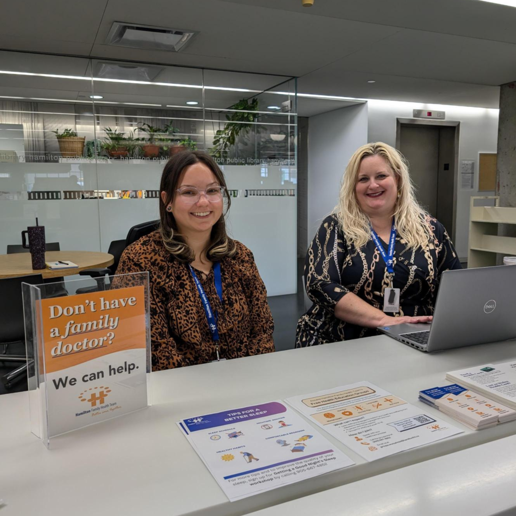 Natasha, a HFHT registered dietitian and Melissa, an HFHT patient care coordinator sit at the Hamilton Central Library Partner Desk and smile at the camera.