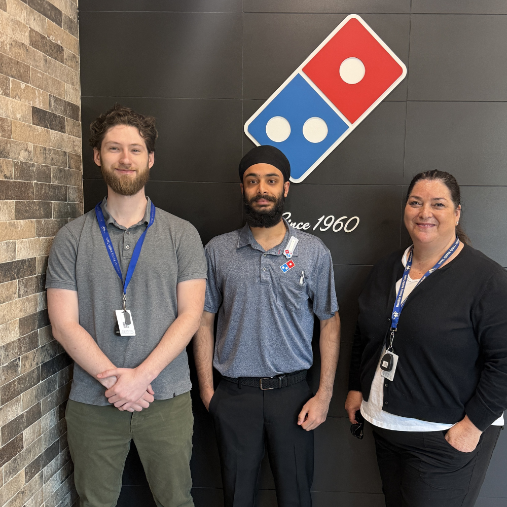 Three people stand and smile at the camera in front of a Dominos Pizza sign.