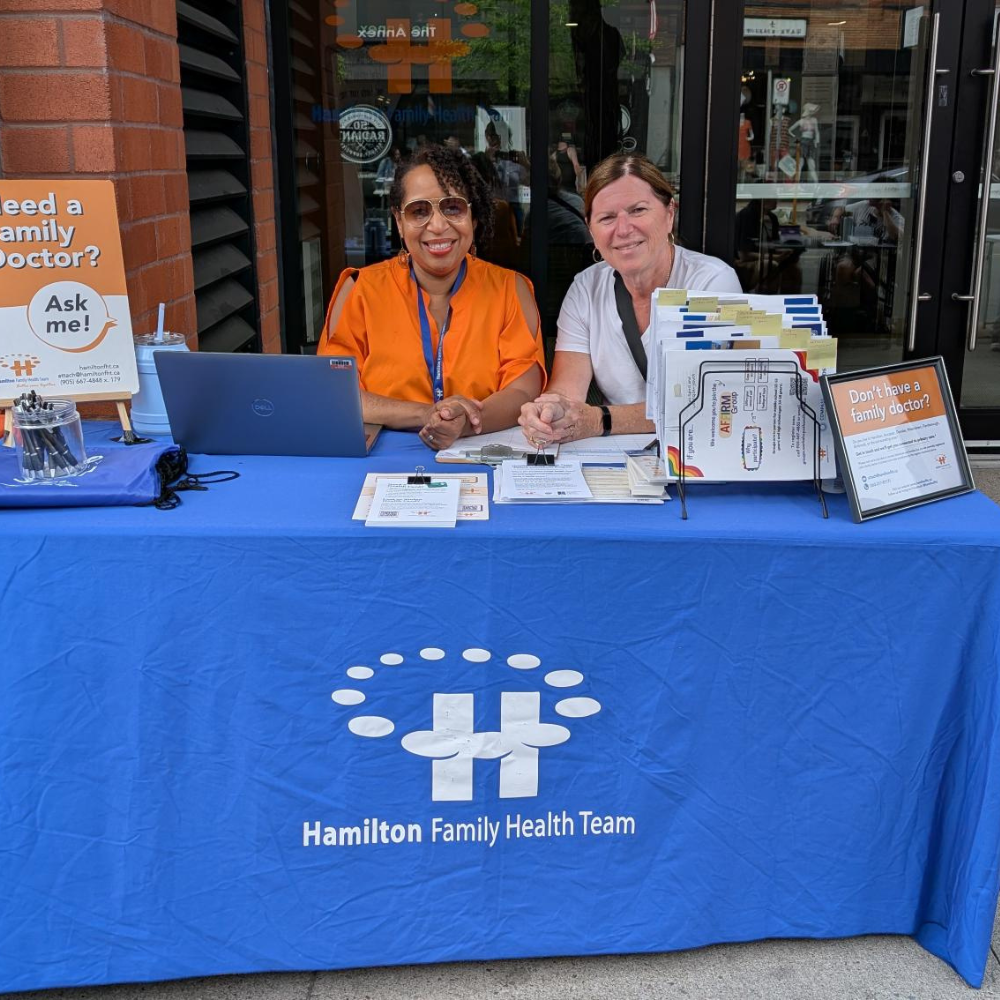 Two HFHT staff members sit outside our home office at 123 James Street at an outreach table during Art Crawl.