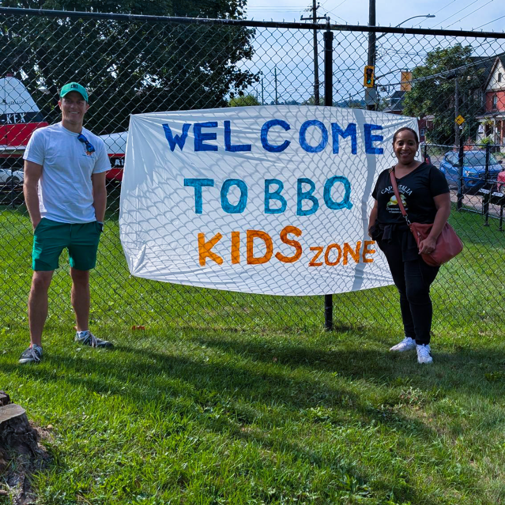 Two HFHT staff members stand beside a sign that says "Welcome to BBQ Kids Zone" at an outreach event at the Eva Rothwell Centre.