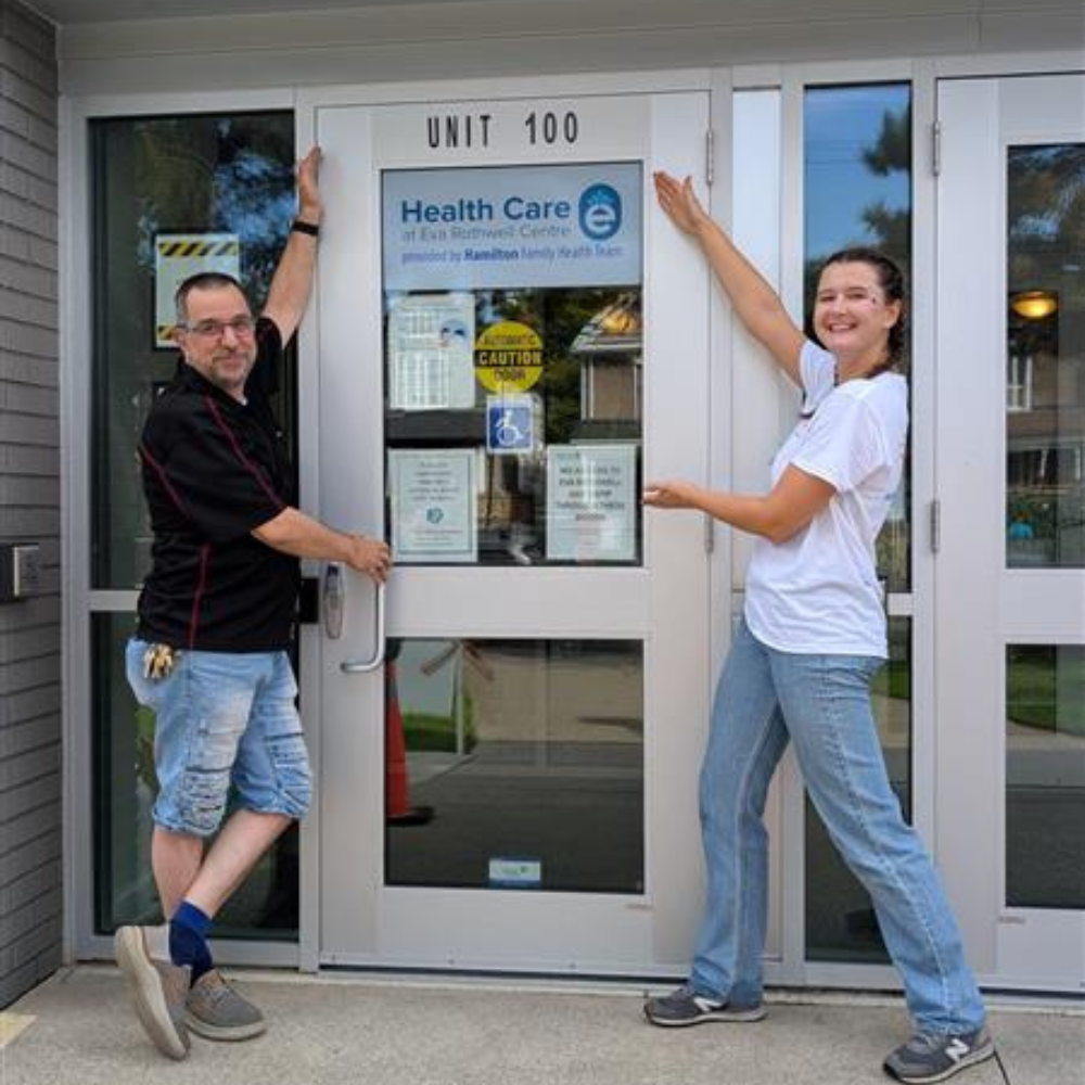 An Eva Rothwell Centre staff member and an HFHT staff member gesture to the "Health Care at Eva Rothwell Centre" sign.
