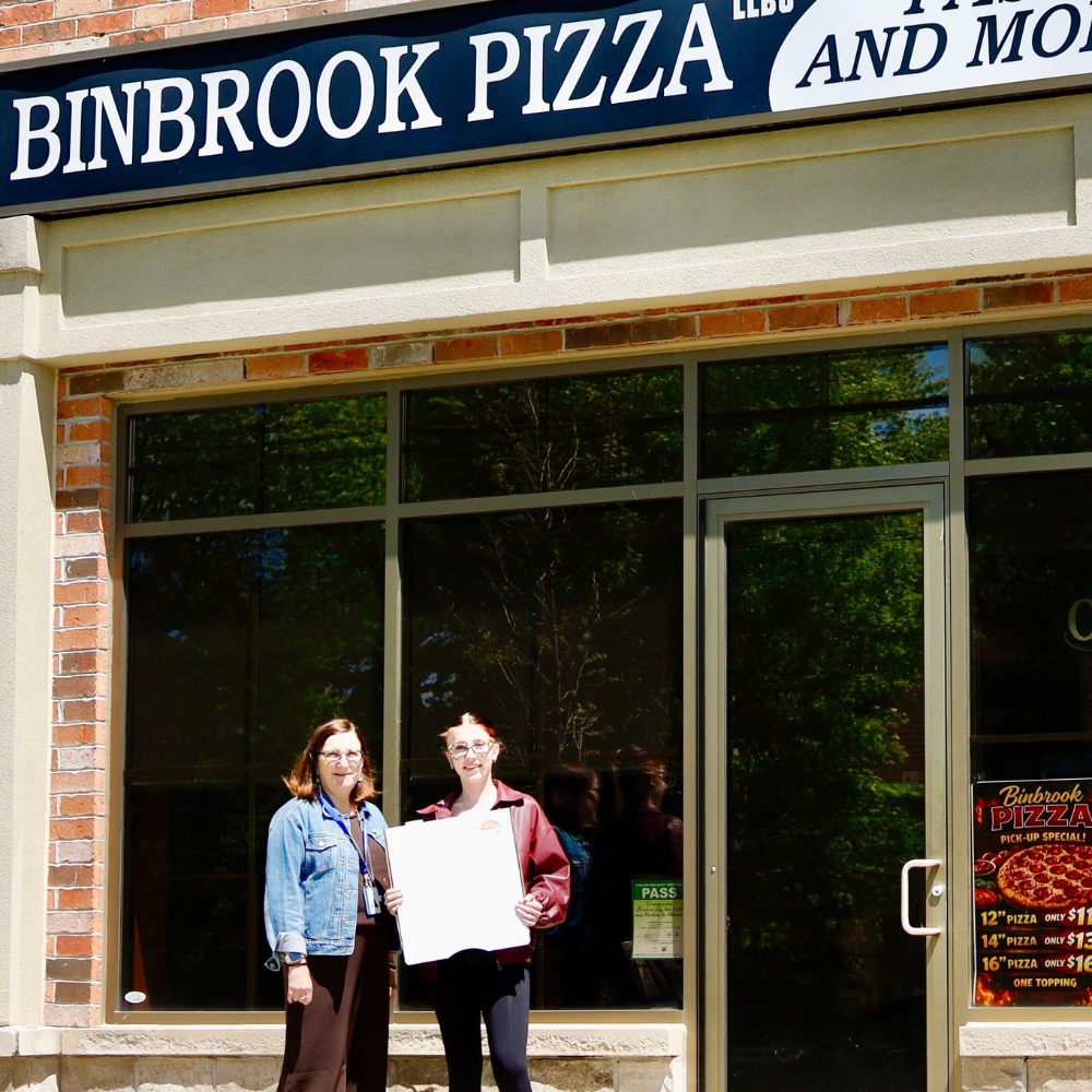 An HFHT Staff member and a Binbrook Pizza staff member stand outside Binbrook Pizza and hold a pizza box.