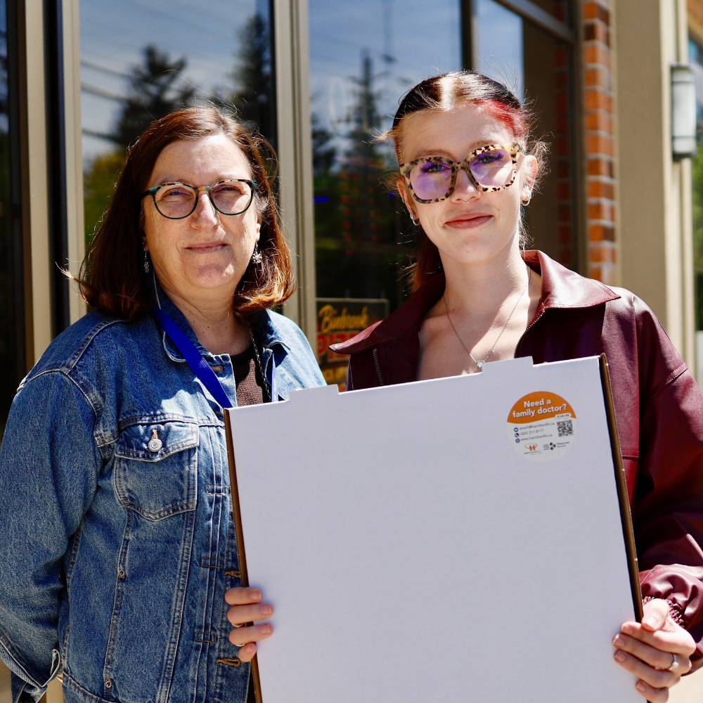 An HFHT Staff member and a Binbrook Pizza staff member stand outside Binbrook Pizza and hold a pizza box. The box has a sticker on it that says "Need a family doctor?"