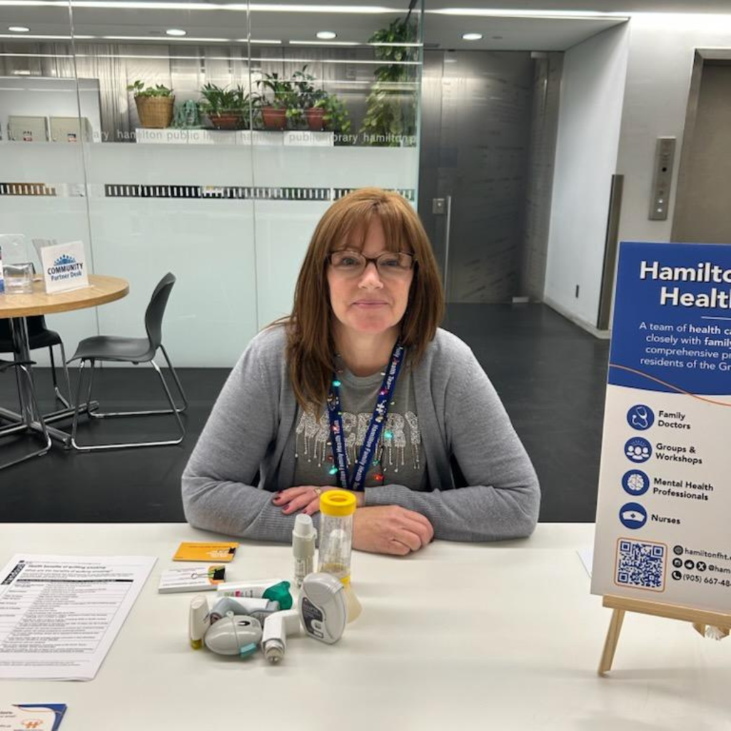 Mary, an HFHT certified respiratory educator, sits at the Hamilton Central Library Partner Desk and smiles at the camera.