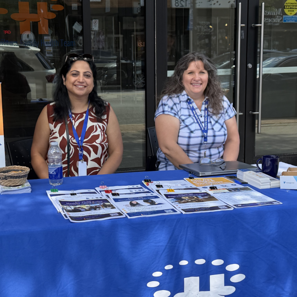 Two HFHT staff members sit outside our home office at 123 James Street at an outreach table during Art Crawl.