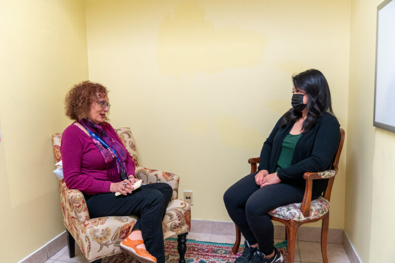 A mental health counsellor and a patient sit face to face in a clinic room. The patient is wearing a mask.