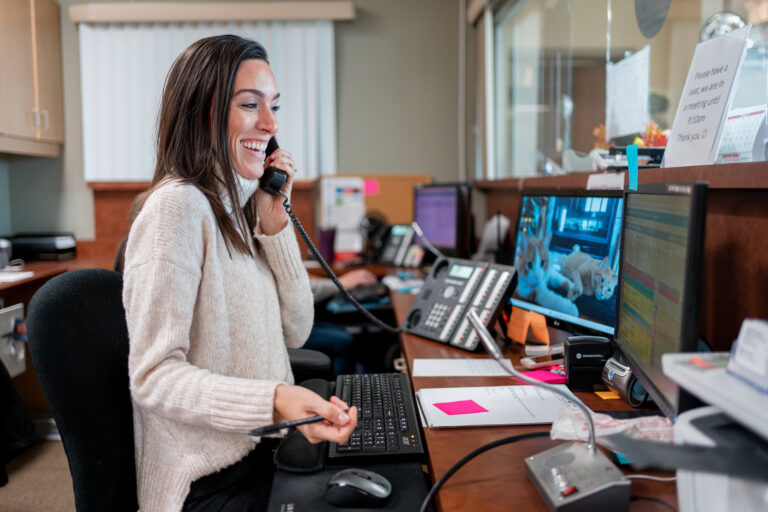 A woman with brown hair sits at a medical reception desk and holds a phone.