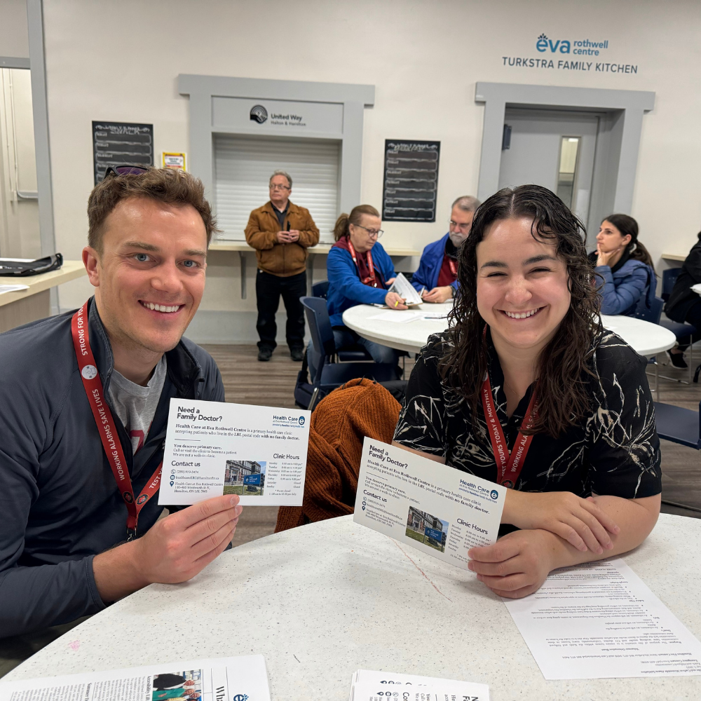 Two HFHT staff members sit inside the Eva Rothwell Centre and hold up flyers that say "Need a family doctor?"