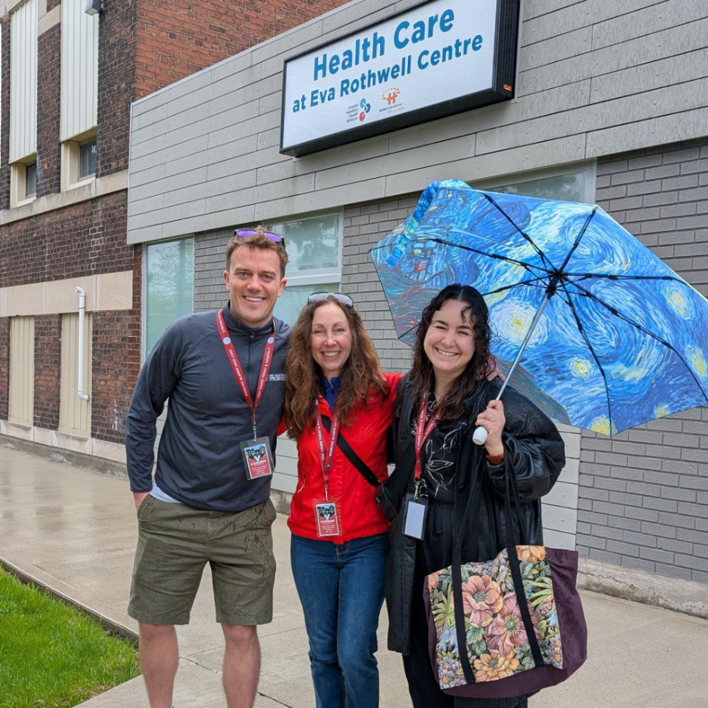 Three HFHT staff members stand outside Health Care at Eva Rothwell Centre. One is holding an umbrella.