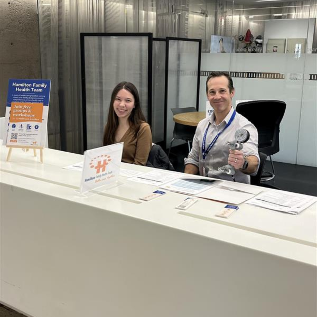 Caroline, a McMaster physiotherapy student, and Jake, an HFHT physiotherapist, sit at the Hamilton Central Library Partner Desk and smile at the camera.