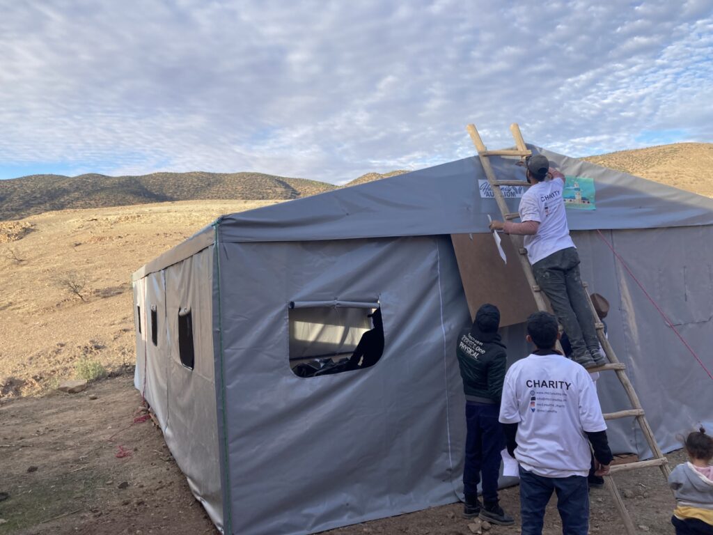 Three people wearing t-shirts that say "Charity" on the back stand next to a grey tent. One person is on a wooden ladder.