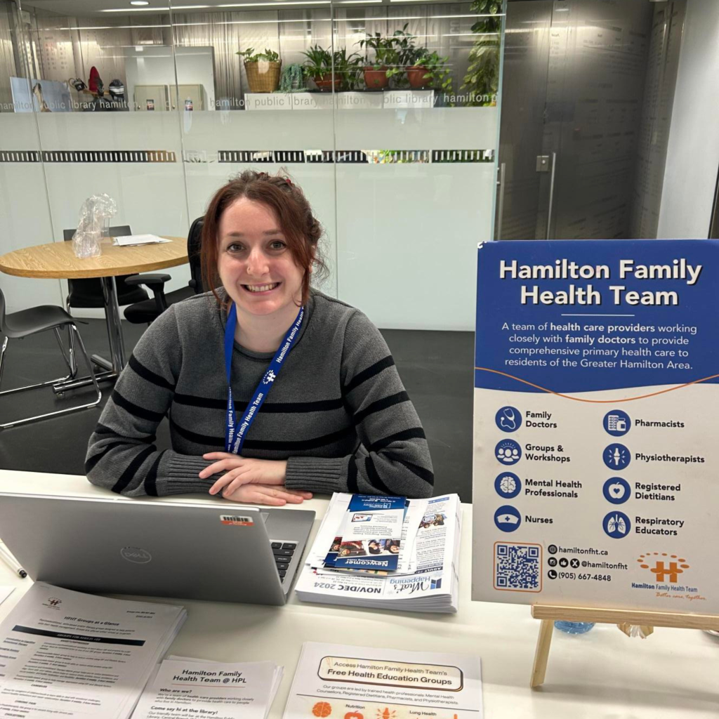 Samm, an HFHT social worker, sits at the Hamilton Central Library Partner Desk and smiles at the camera.