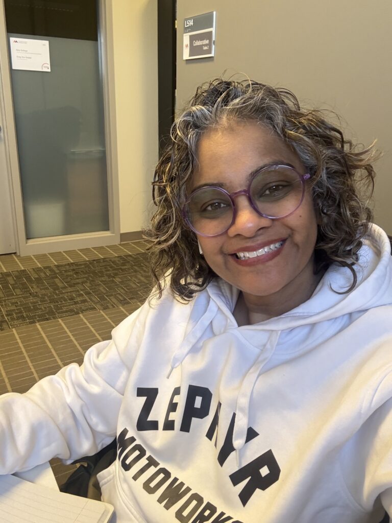 A woman with glasses and short curly hair wears a white hoodie and smiles at the camera.