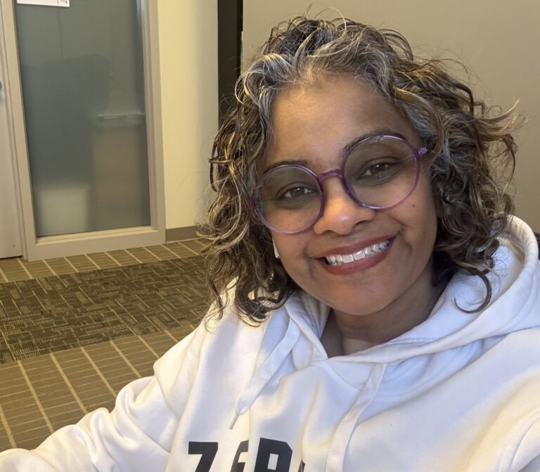 A woman with glasses and short curly hair wears a white hoodie and smiles at the camera.