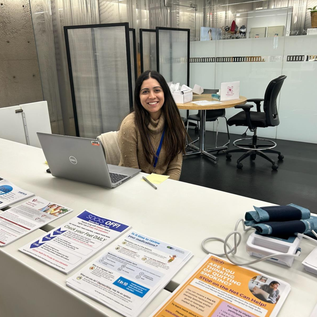Baldeep, an HFHT pharmacist, sits at the Hamilton Central Library Partner Desk and smiles at the camera.