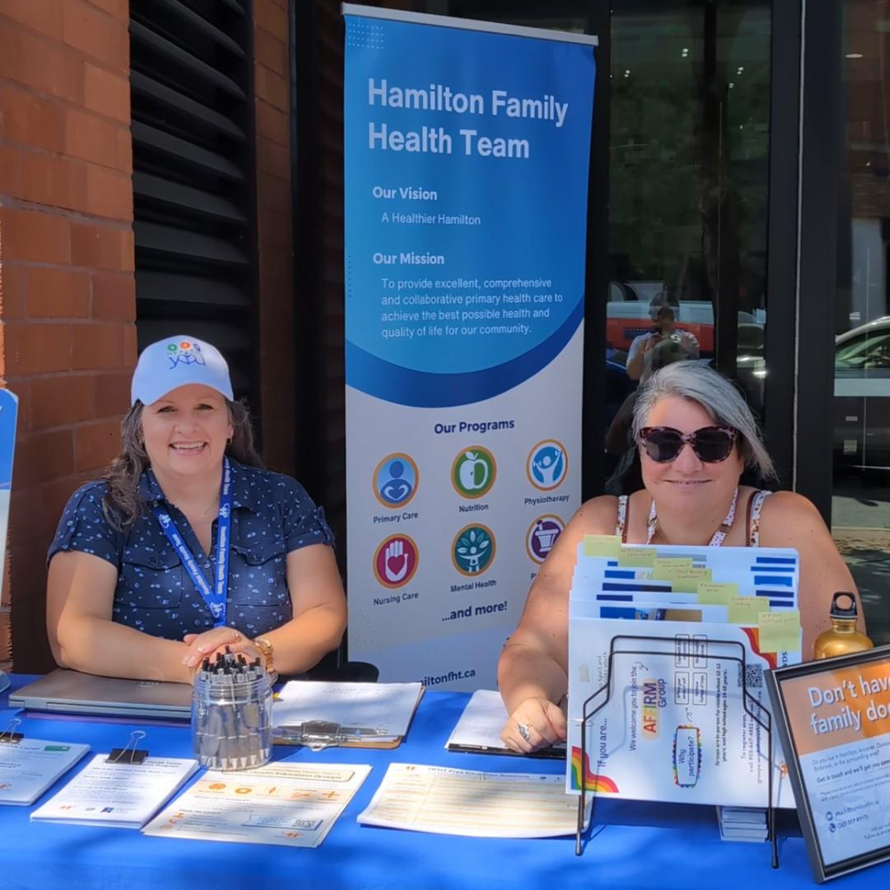 Two HFHT staff members sit outside our home office at 123 James Street at an outreach table during Art Crawl.