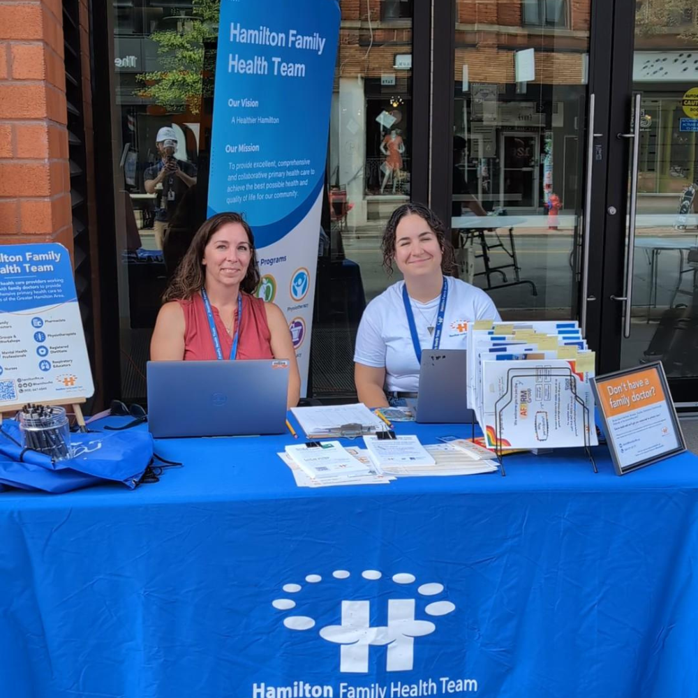 Two HFHT staff members sit outside our home office at 123 James Street at an outreach table during Art Crawl.