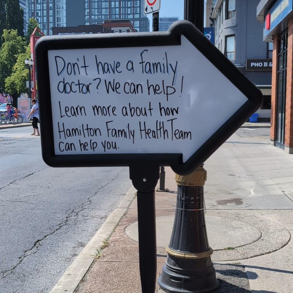 A whiteboard sign that says "Don't have a family doctor? We can help! Learn more about how Hamilton Family Health Team can help you."