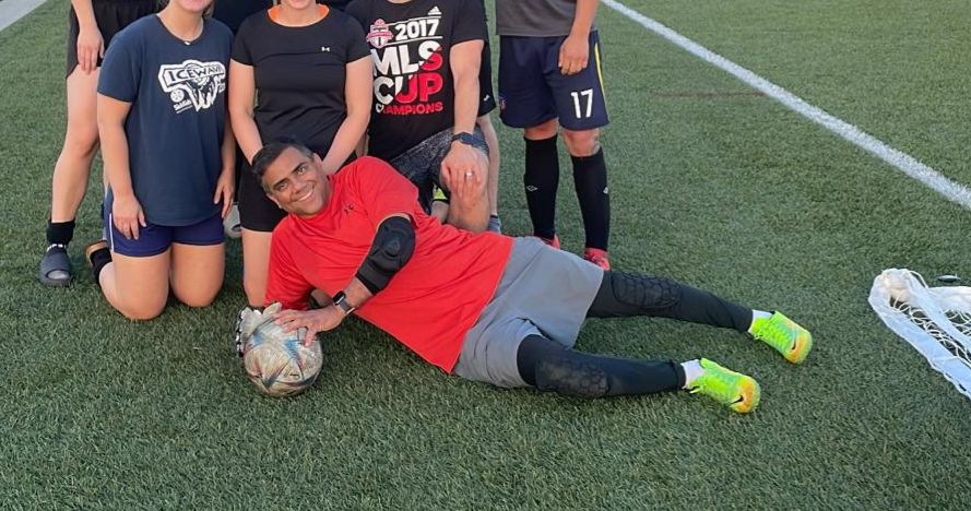 A group of people on a soccer field after a soccer game. At the front a man holds a soccer ball, wearing a red shirt, grey shorts and neon green running shoes, and smiles at the camera.