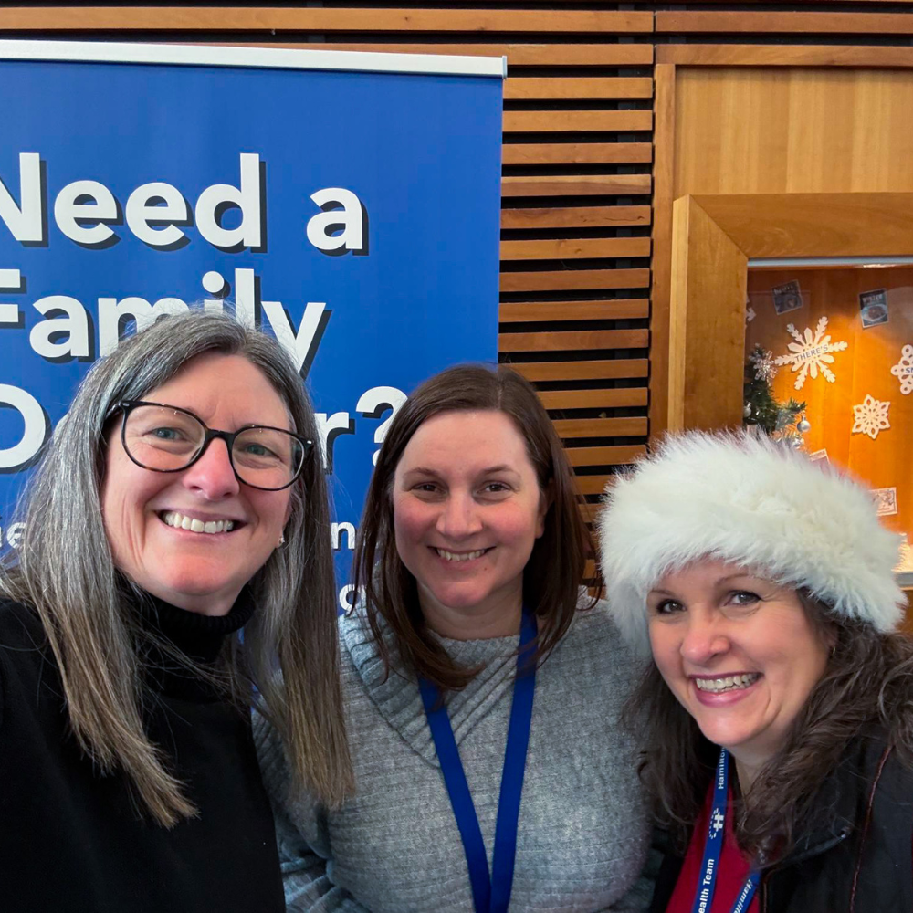 Three HFHT staff members smile at the camera at an outreach event. One person is wearing a Santa hat.