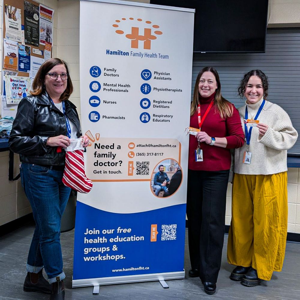 Three HFHT staff members smile at the camera while standing next to an HFHT banner at an outreach event. One person is holding a Christmas stocking.