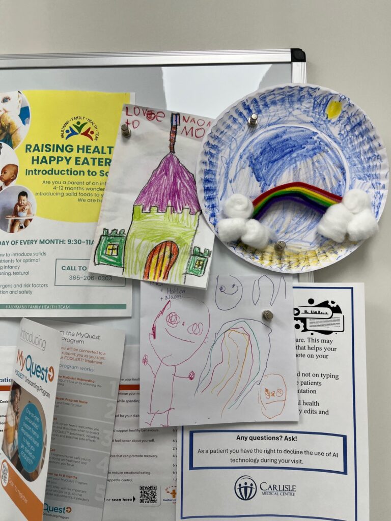 Three pieces of children's art, of a house, people, and a rainbow, are hung by magnets on a whiteboard along with medical flyers.