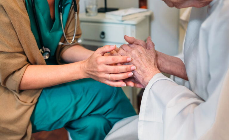A doctor wearing teal scrubs and a stethoscope holds the hands of an elderly patient.
