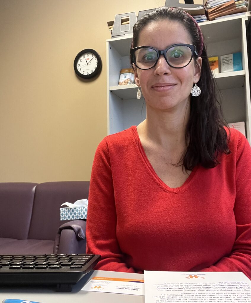 A woman with shoulder-length brown hair sits at a desk and smiles at the camera. She is wearing a bright red sweater, silver earrings, and black framed glasses.