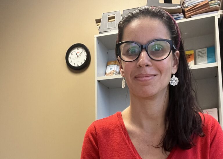 A woman with shoulder-length brown hair sits at a desk and smiles at the camera. She is wearing a bright red sweater, silver earrings, and black framed glasses.
