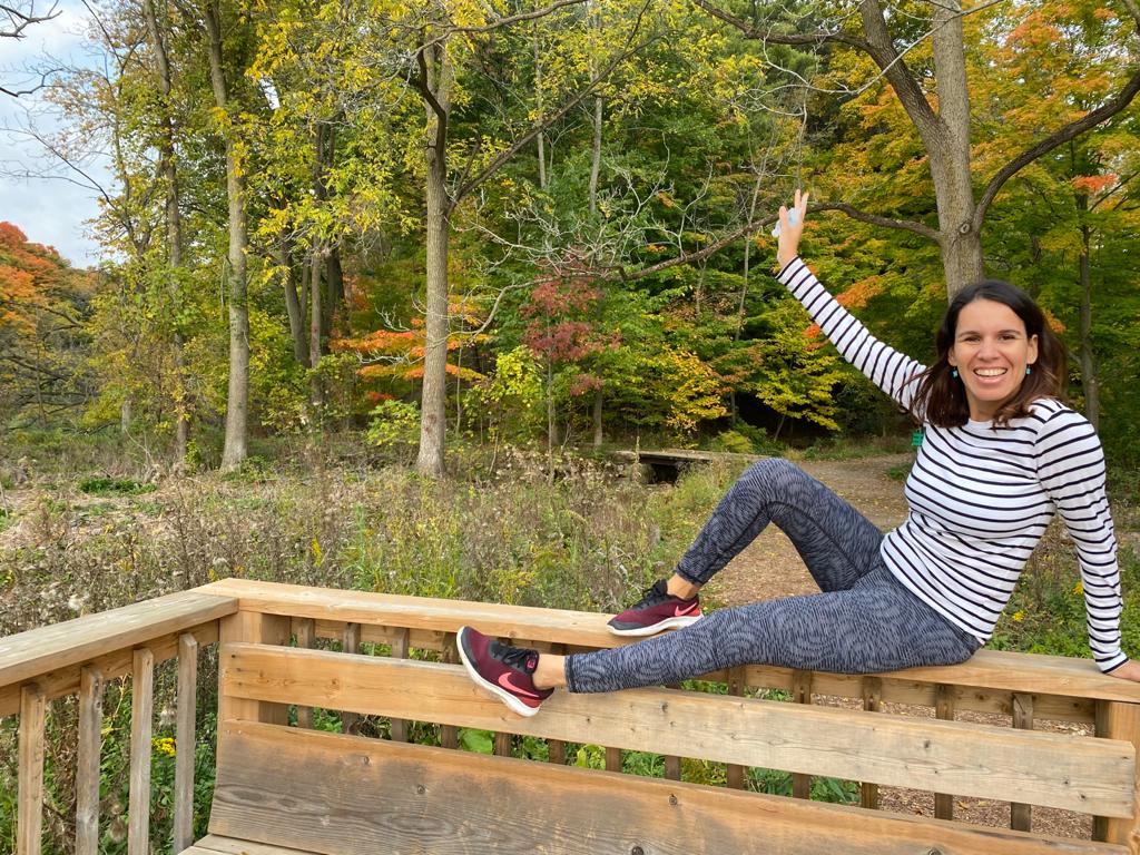A woman sits on a wooden fence with one hand in the air and smiles at the camera. She is surrounded by trees in the fall. She's wearing a white and black striped shirt, grey leggings, and maroon nike sneakers.