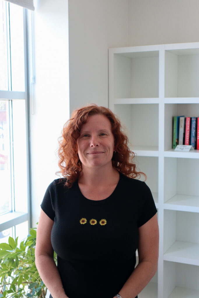 A woman with red curly hair wears a black t-shirt and stands in front of a white bookshelf and window. She is smiling at the camera.