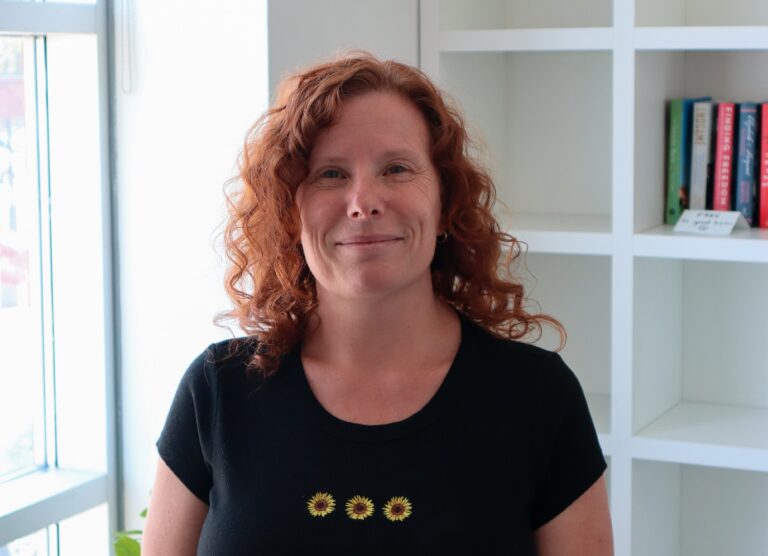 A woman with red curly hair wears a black t-shirt and stands in front of a white bookshelf and window. She is smiling at the camera.