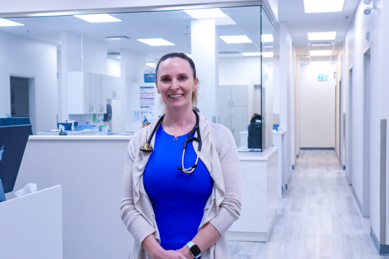 A doctor stands inside a healthcare clinic and smiles at the camera. She's wearing a stethescope, a blue top, a white cardigan, and has her hair pulled back.