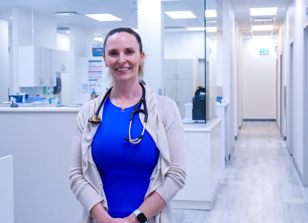 A doctor stands inside a healthcare clinic and smiles at the camera. She's wearing a stethescope, a blue top, a white cardigan, and has her hair pulled back.