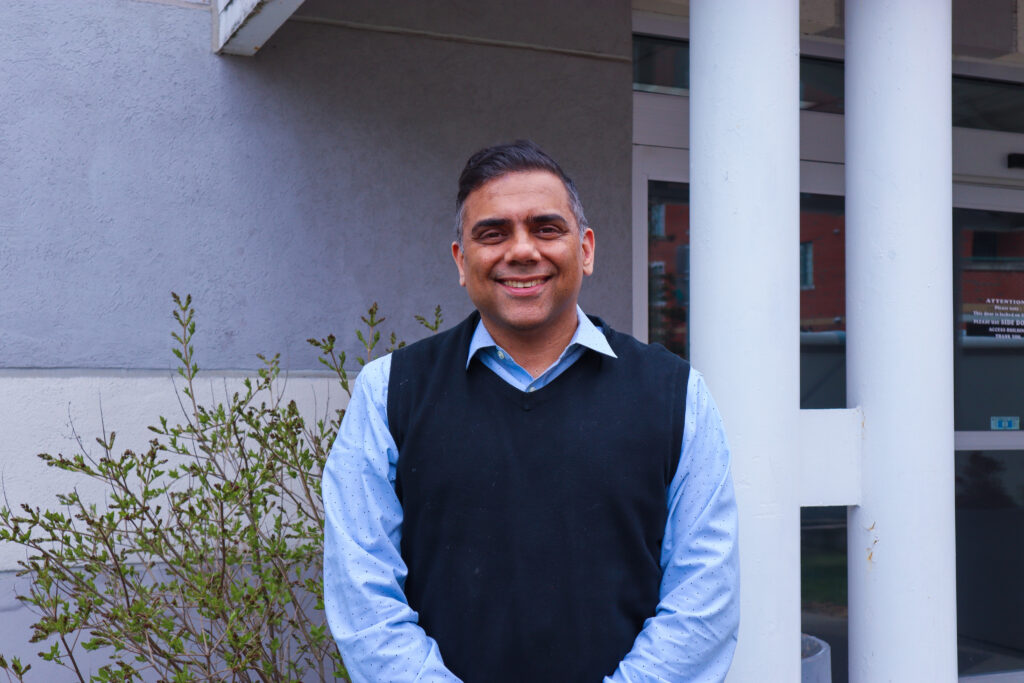 Dr. Mustafa Warsi stands outside his clinic and smiles at the camera. He is wearing a light blue collared shirt and a dark blue sweater vest.