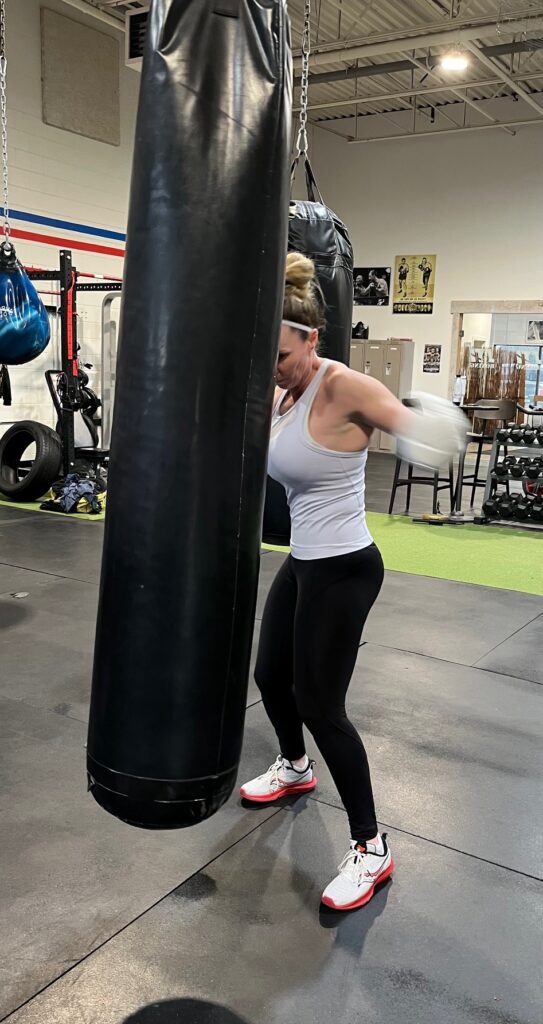 A woman wearing a white tank top, black leggings, running shoes and white boxing gloves hits a punching bag in a boxing gym.