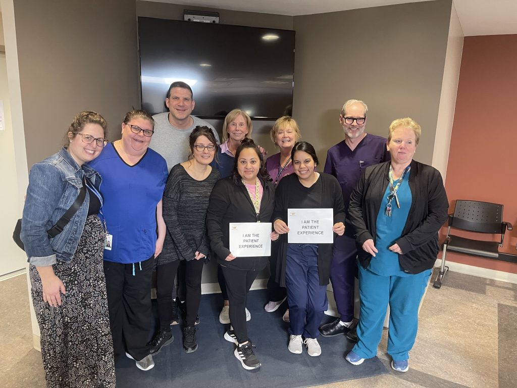 Harvard Square Family Physicians Practice Team hold "I am the patient experience" signs and smile at the camera.