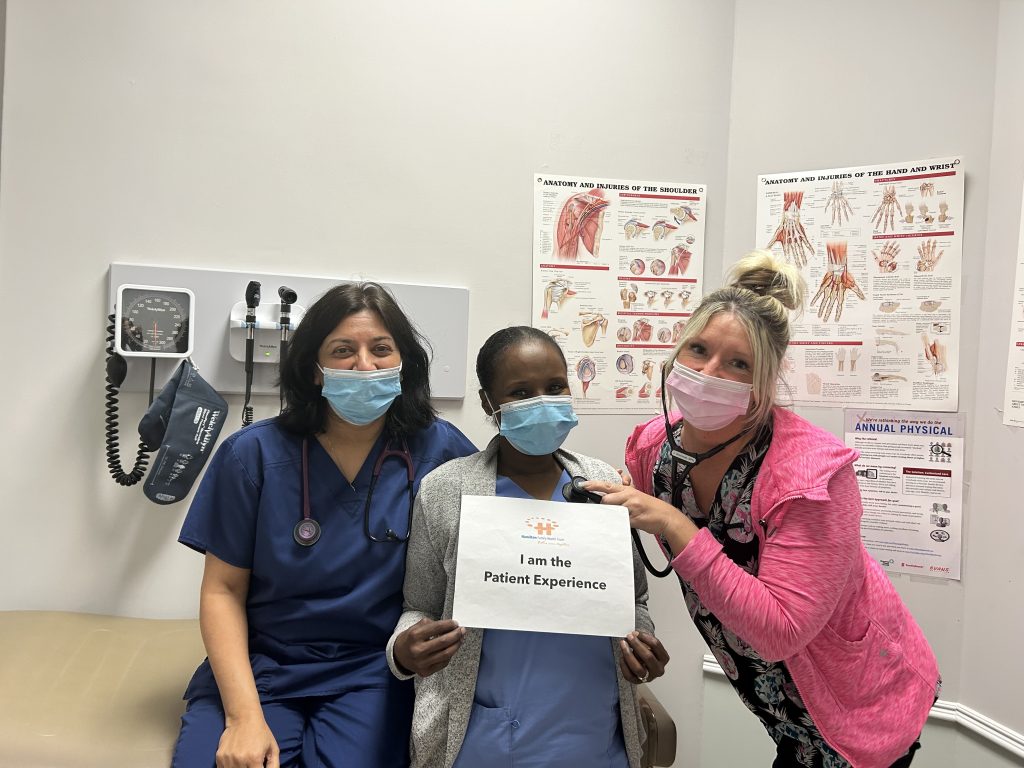 Drs. Ghelani and Nathoo’s
Practice Team hold an "I am the patient experience sign" while wearing masks in a clinical exam room.