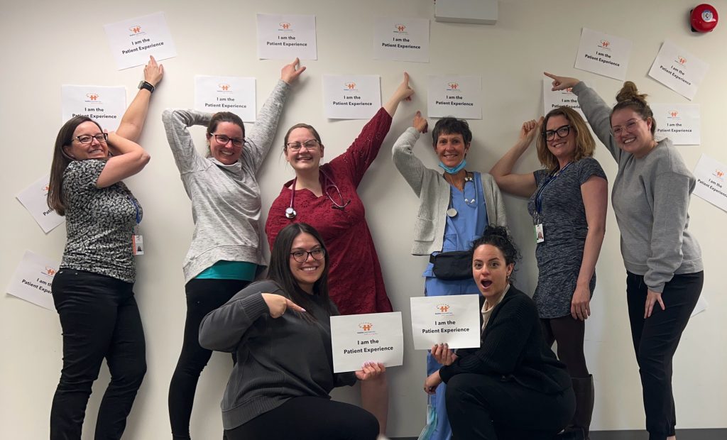 Queensdale Hub practice team pointing to many "I am the patient experience" signs taped to the wall.