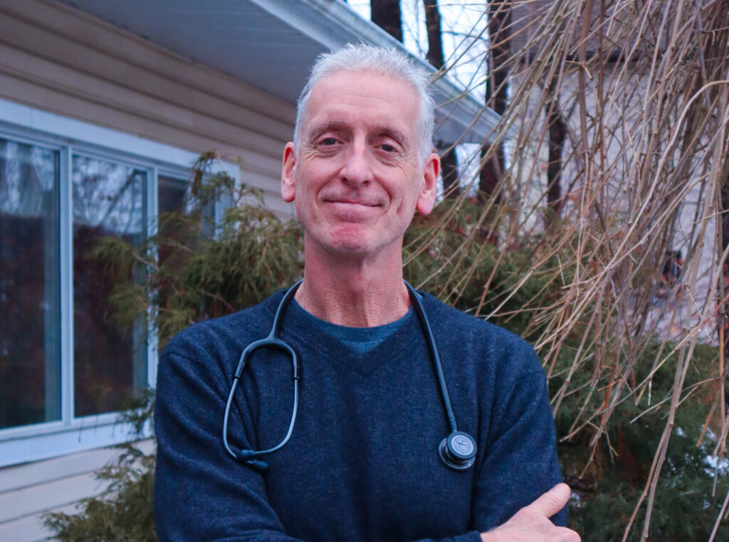 A man with white hair smiles at the camera while he stands outside a healthcare clinic. He is wearing a blue sweater and a stethoscope.