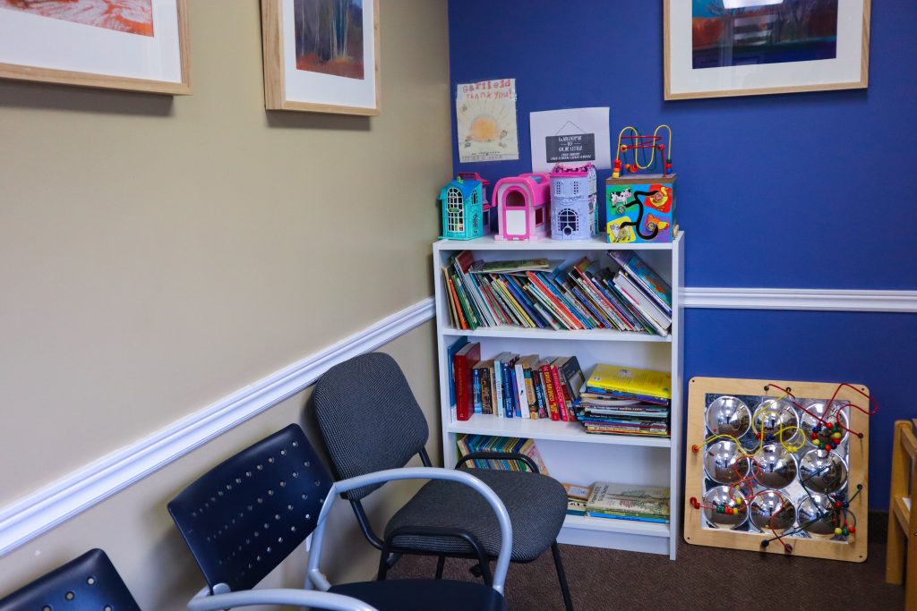 A corner of a healthcare clinic with chairs, a bookshelf, and toys for children. Art is hung on the walls.