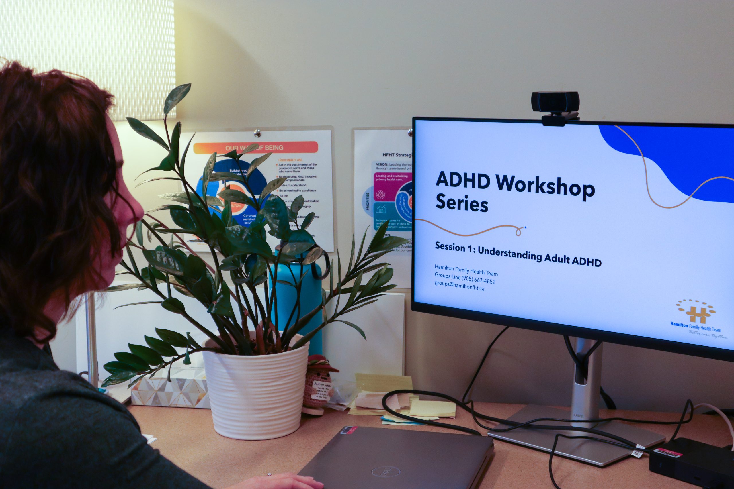 A person. sitting at a desk with a plant, looking at a desktop computer, with a presentation on ADHD for Adults.
