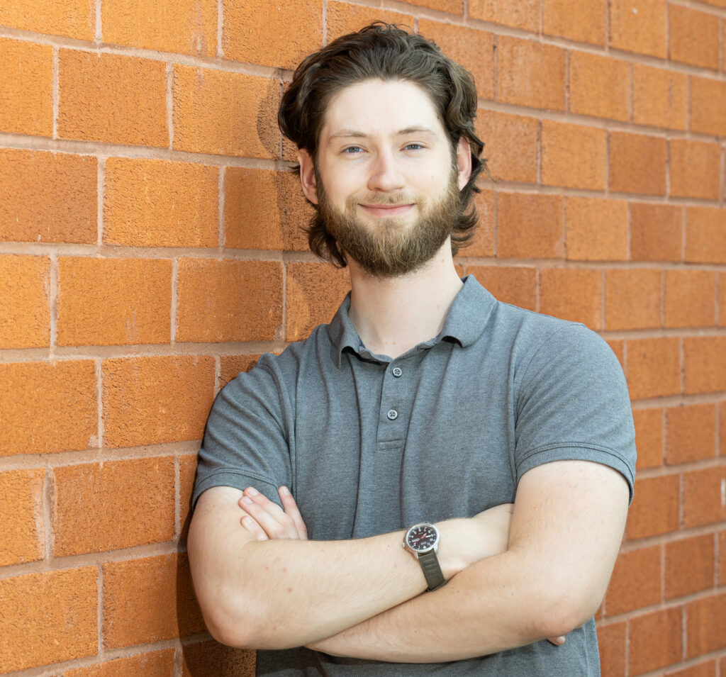 Jacob Pirgorsch by a brick wall, arms crossed