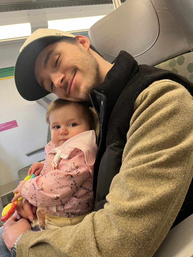 A man wearing a black vest, tan sweater, and baseball cap holds his young daughter who is wearing a pink coat with unicorns. They are sitting on a train.