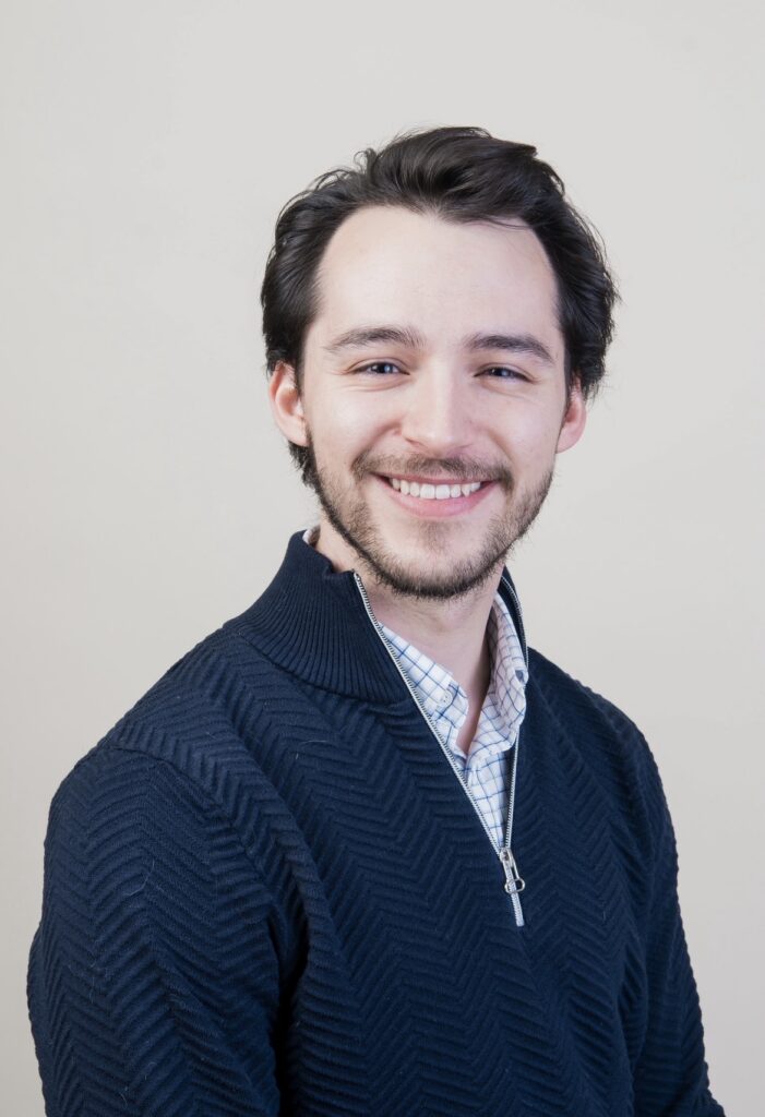 A man with short brown hair and a short beard smiles at the camera. He's wearing a dark blue sweater and a collared shirt.