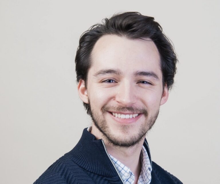 A man with short brown hair and a short beard smiles at the camera. He's wearing a dark blue sweater and a collared shirt.