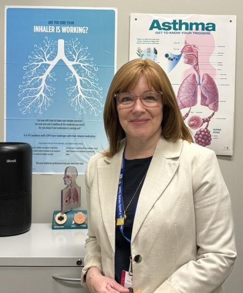 A woman with straight red-brown hair and glasses smiles at the camera. She is standing in front of educational posters about asthma. She is wearing a white blazer and black shirt.