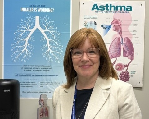 A woman with straight red-brown hair and glasses smiles at the camera. She is standing in front of educational posters about asthma. She is wearing a white blazer and black shirt.