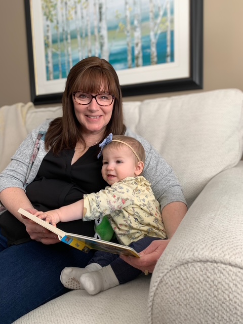 A woman with straight red-brown hair and glasses holds a small child and smiles at the camera.