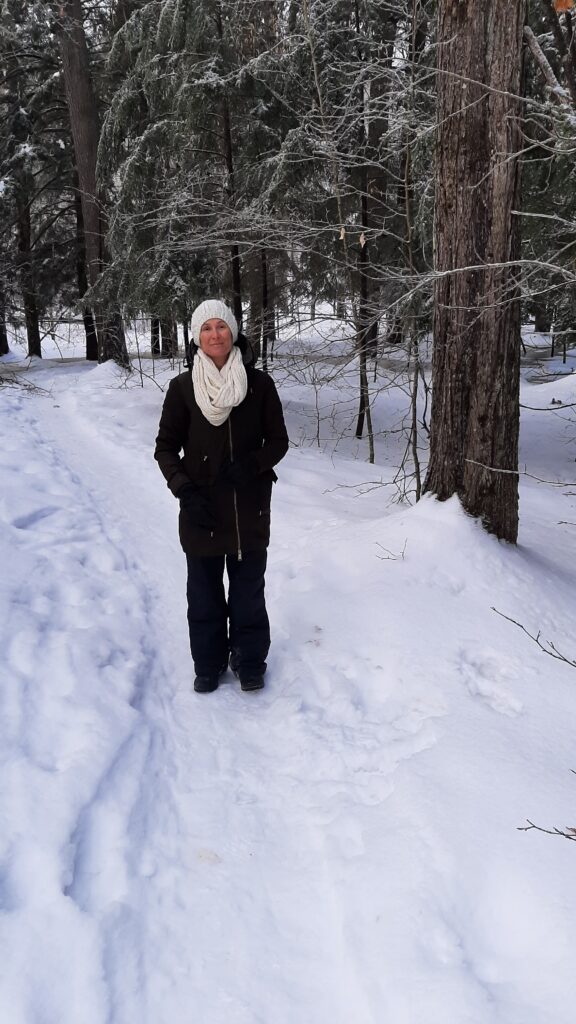 A woman in a black coat, snowpants and boots and a white scarf and hat stands in the snowy forest.